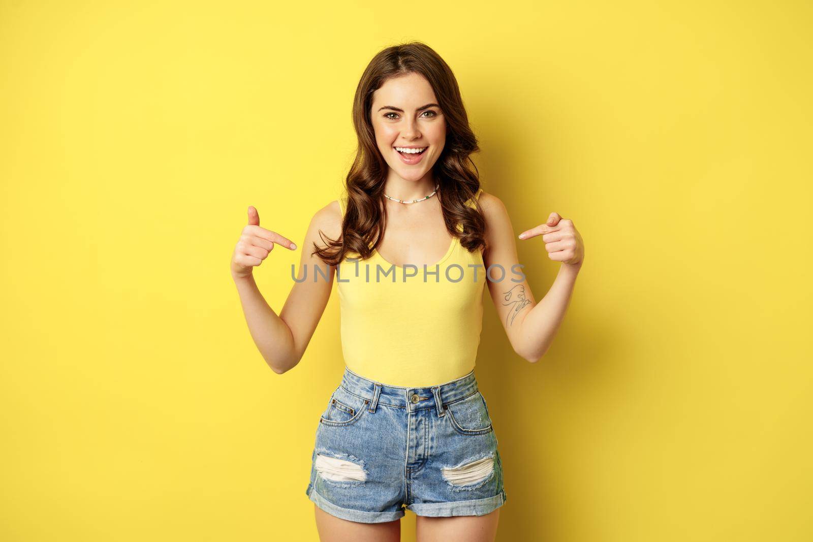 Happy attractive woman pointing fingers at herself, self-promoting, something personal, smiling and looking proud or confident, standing over yellow background.