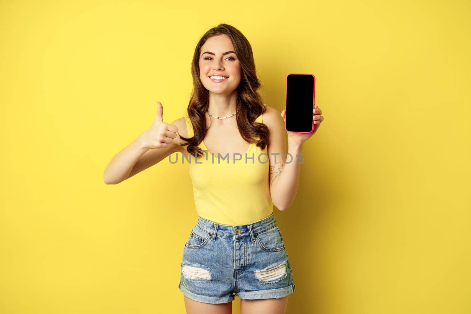 Happy smiling stylish girl showing thumbs up and smartphone app interface, mobile phone display, satisfied with website, online shopping application, standing over yellow background.