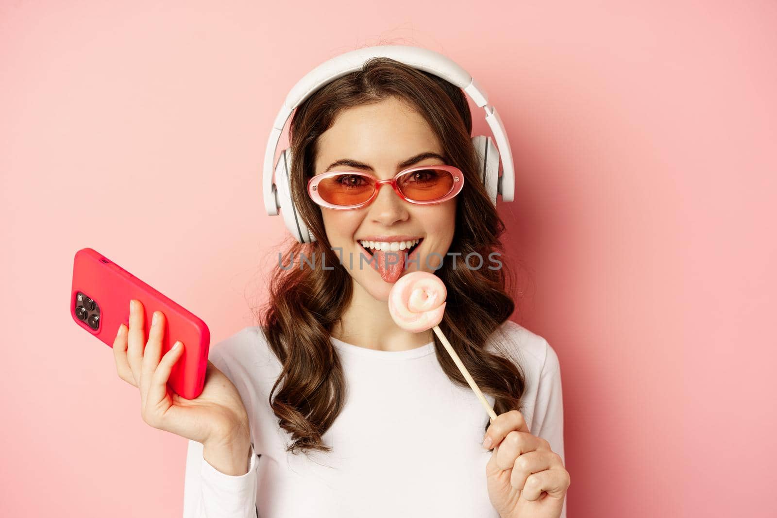 Close up portrait of stylish glamour girl, beautiful woman in headphones, listening music with smartphone, wearing sunglasses and licking lolipop.