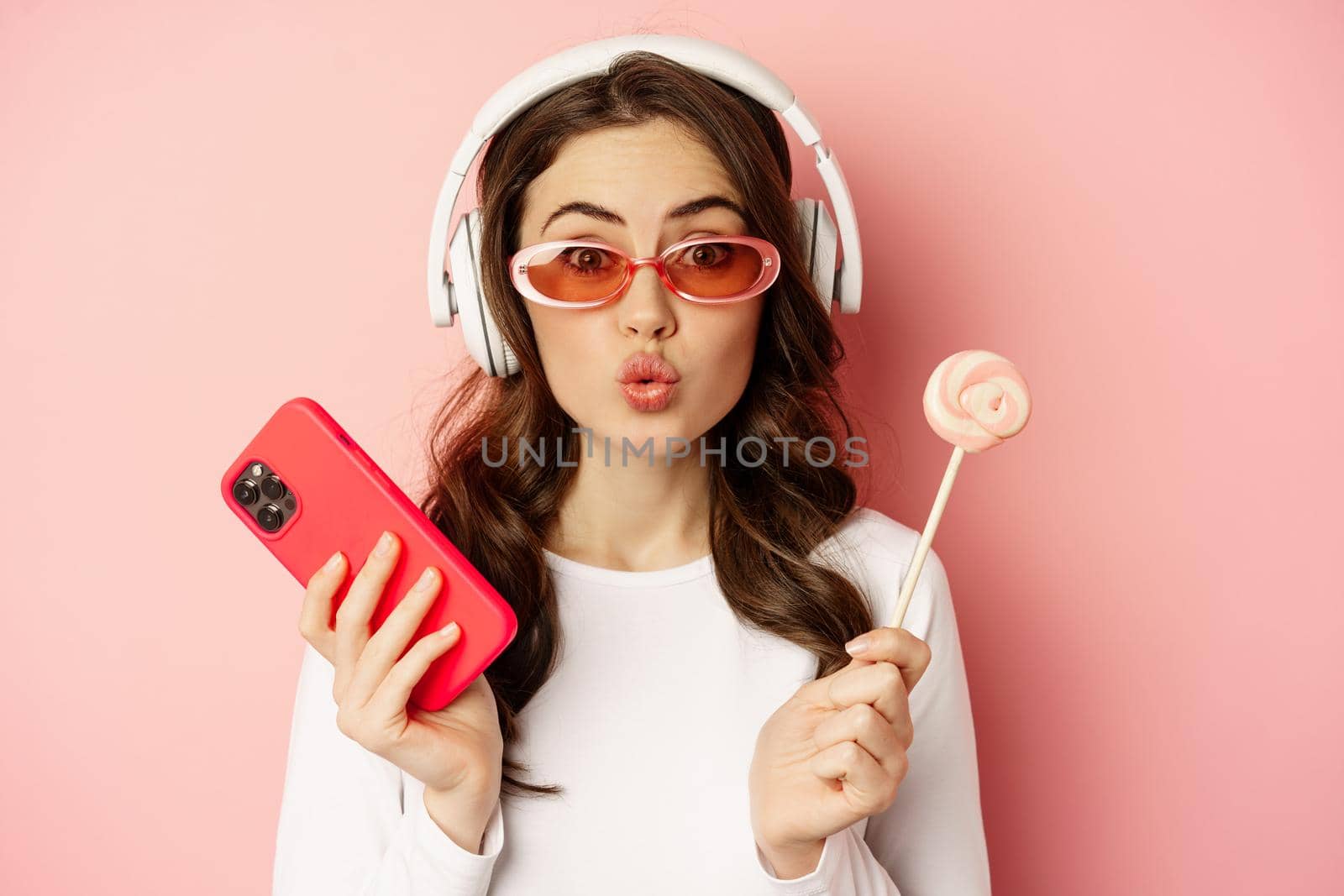 Stylish young woman with lolipop and cellphone, wearing sunglasses and headphones, listening music, standing over pink background.
