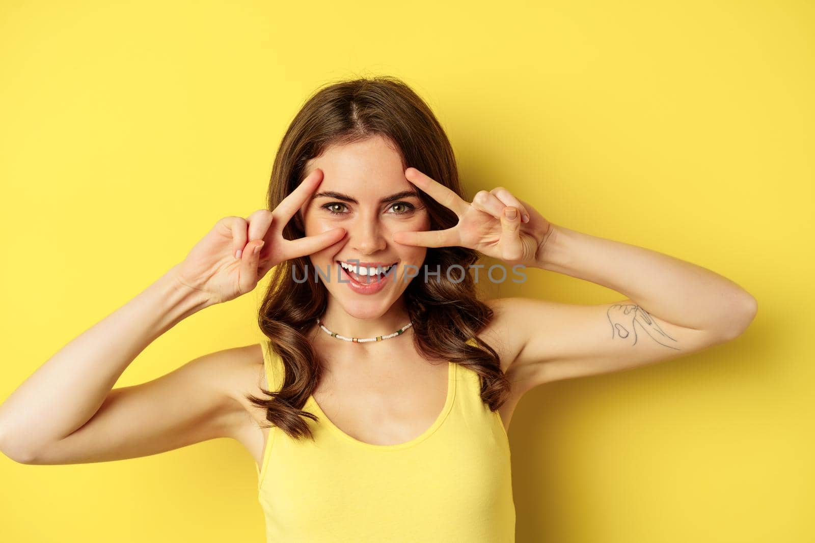 Close up portrait of smiling beautiful girl, showing peace v-sign and looking happy, posing for summer photo, standing against yellow background.