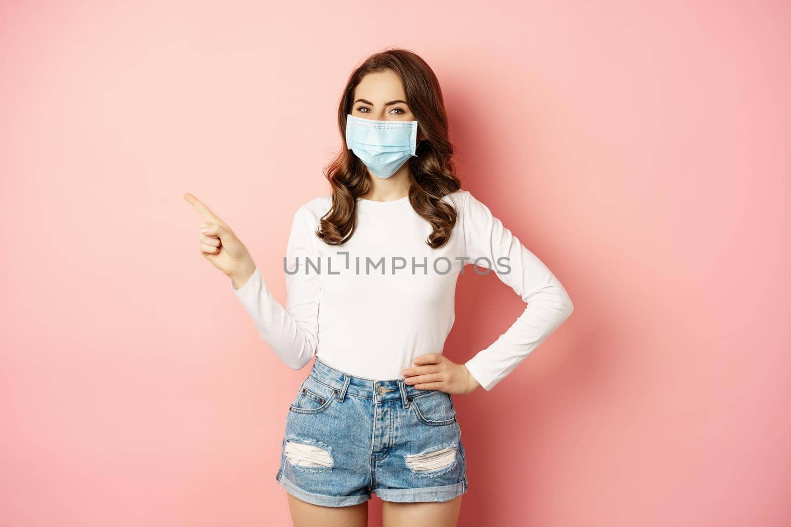 Beautiful stylish woman in medical face mask from covid, pointing finger at upper left corner, showing advertisement, standing over pink background.