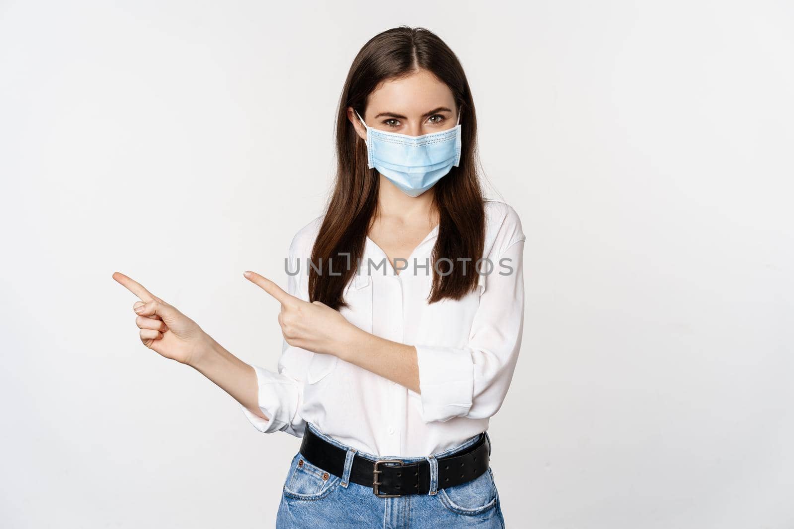 Portrait of corporate woman in face medical mask, pointing fingers left, showing logo, company banner, standing against white background.