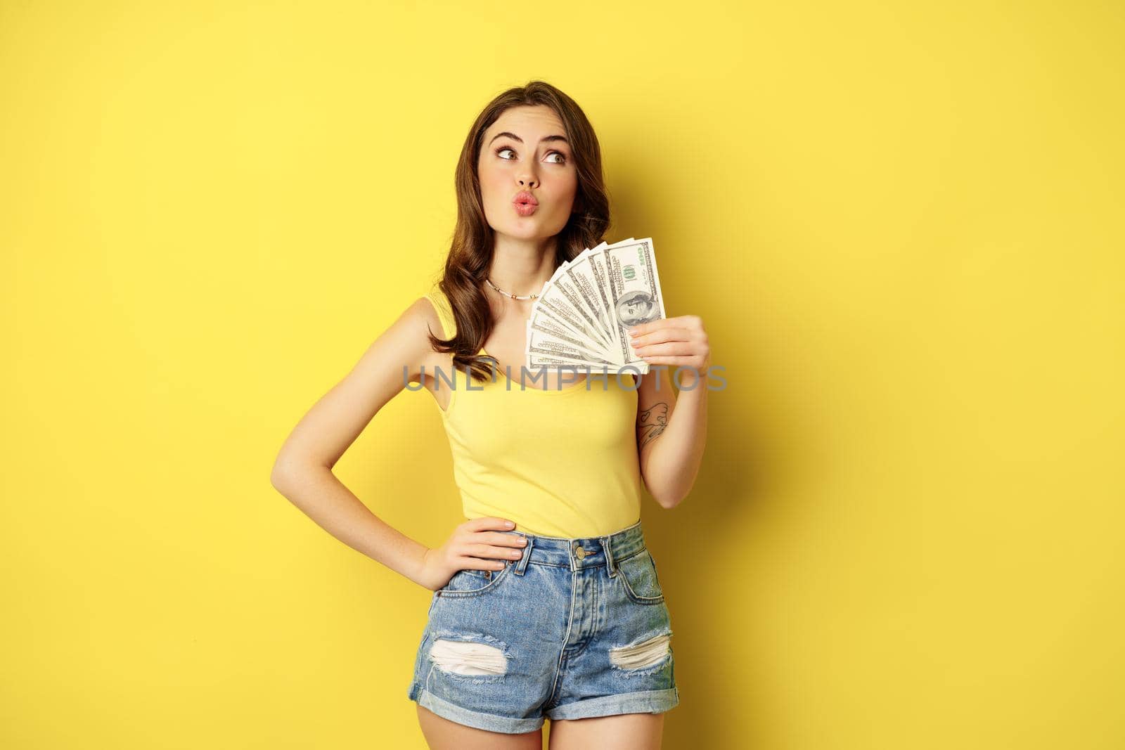 Shopping, credit and money concept. Young brunette woman showing cash and smiling pleased, standing over yellow background by Benzoix
