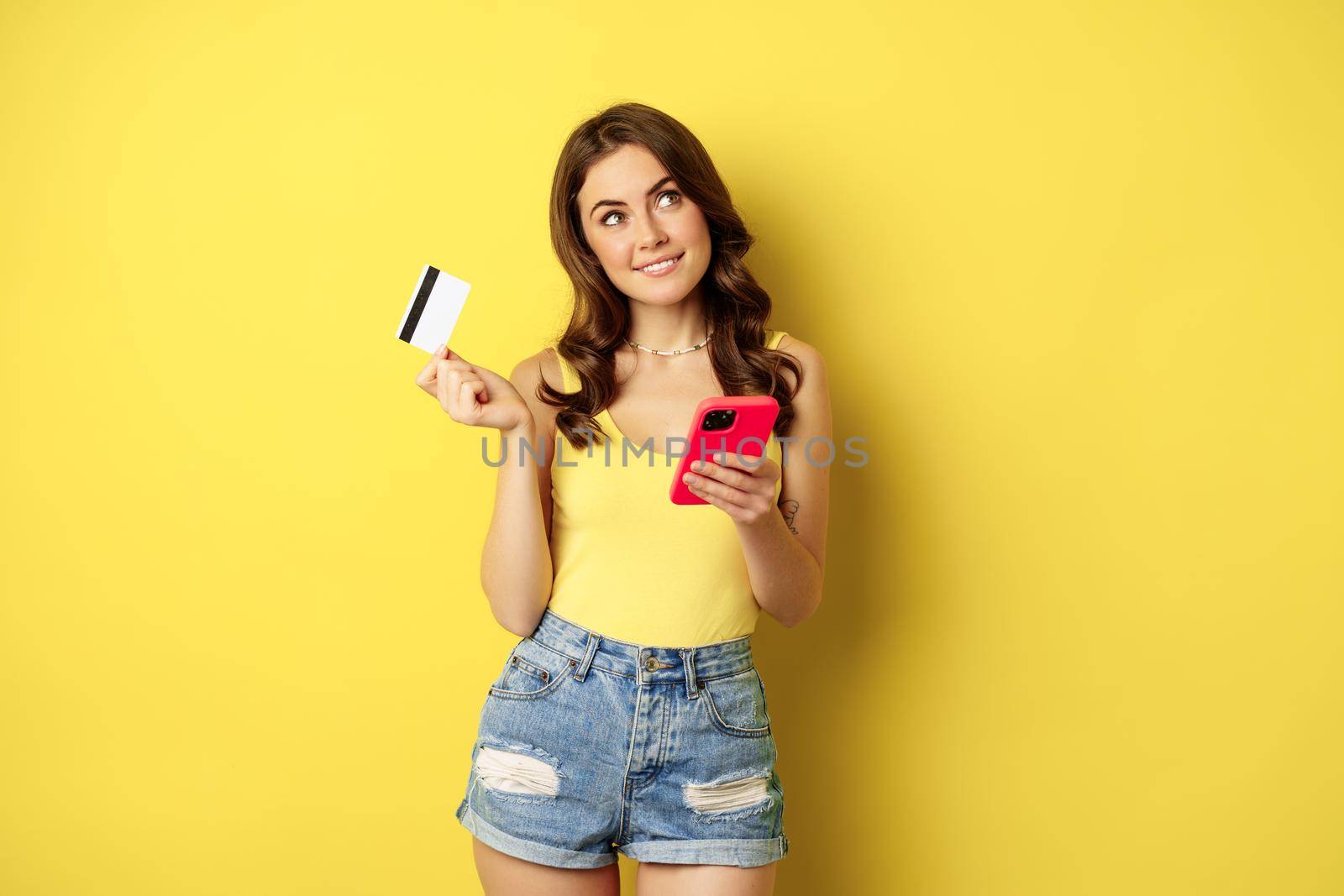 Online shopping. Stylish brunette woman holding smartphone and credit card, paying in app, using mobile phone application, buying smth, standing over yellow background.
