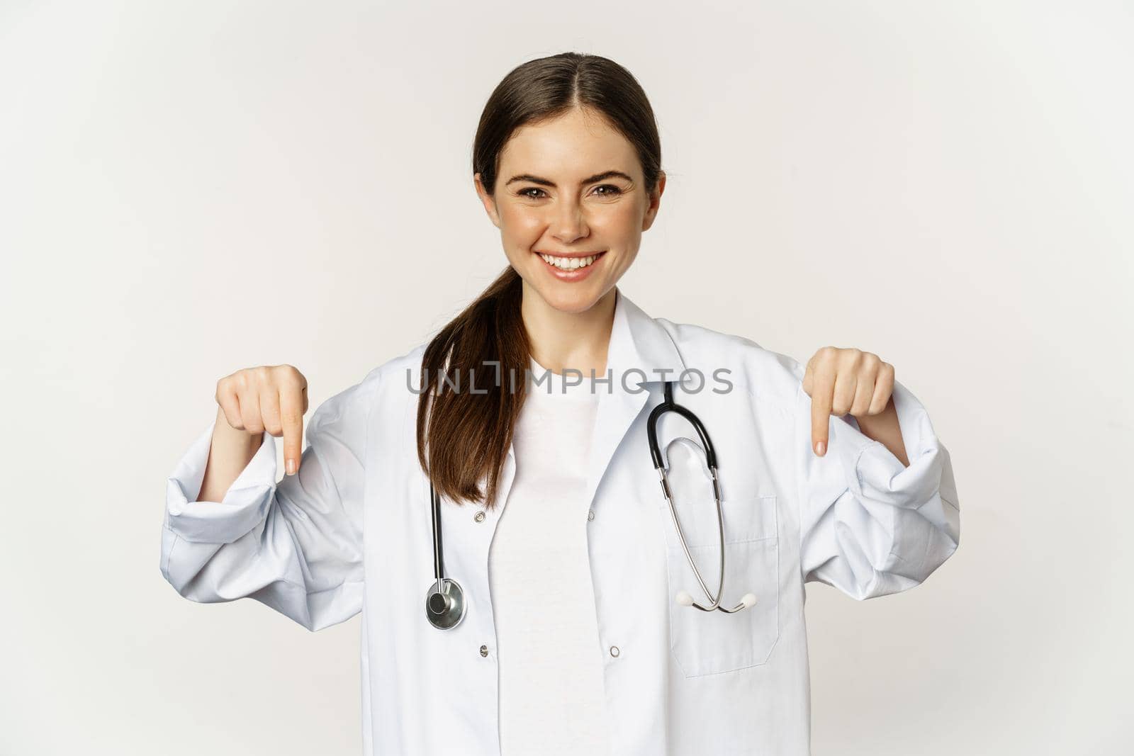 Portrait of happy female doctor, pointing fingers down and smiling, demonstrating promo offer, discount, standing over white background.