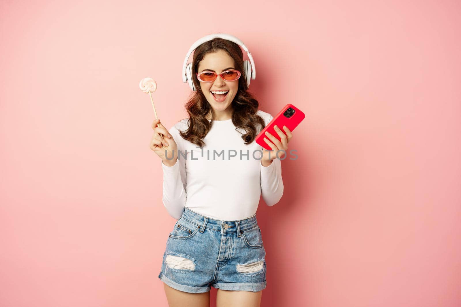 Dancing woman in sunglasses, listening music in headphones, holding lolipop and smartphone, laughing and smiling happy, standing over pink background.