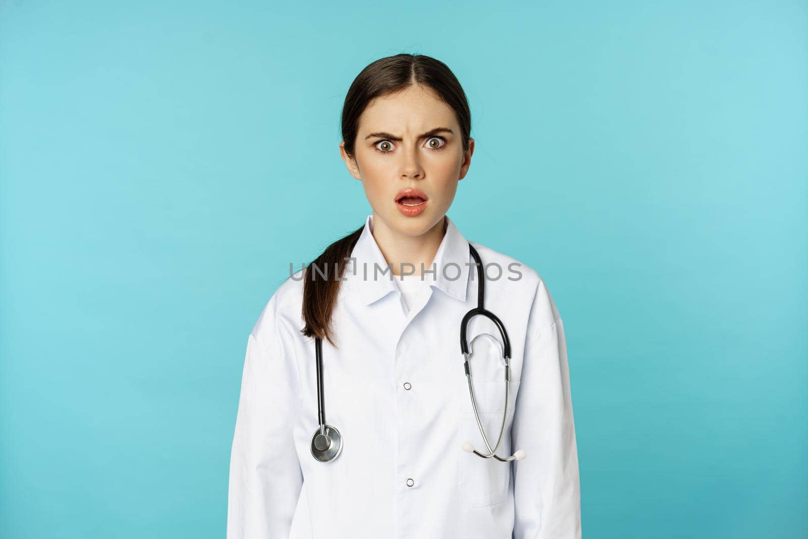Portrait of shocked woman doctor, female hospital intern in white coat, looking concerned and confused at camera, disbelief face, standing over torquoise background.