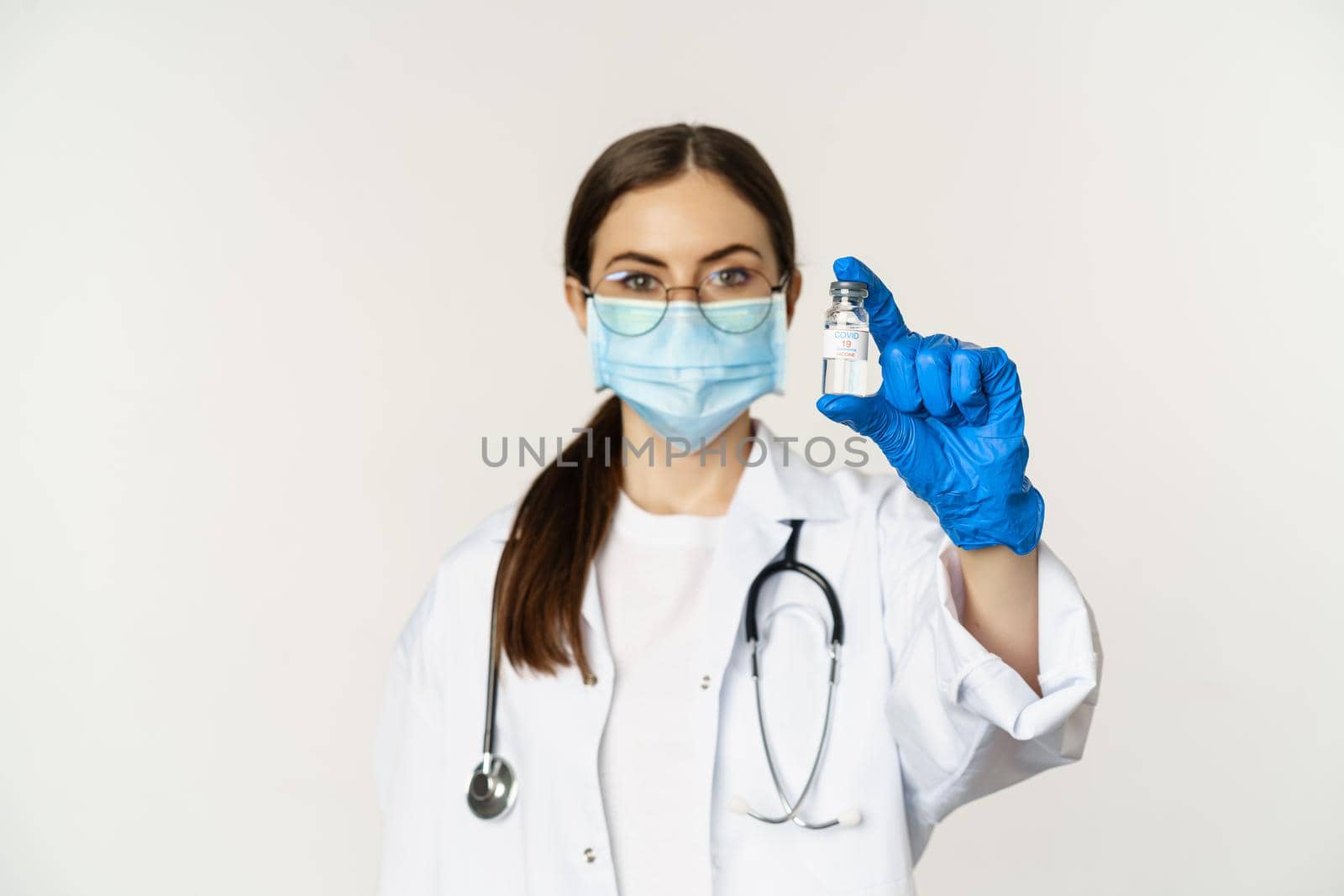 Portrait of young woman, doctor in medical face mask and uniform, showing vaccine, covid-19 vaccination campaign, standing over white background.