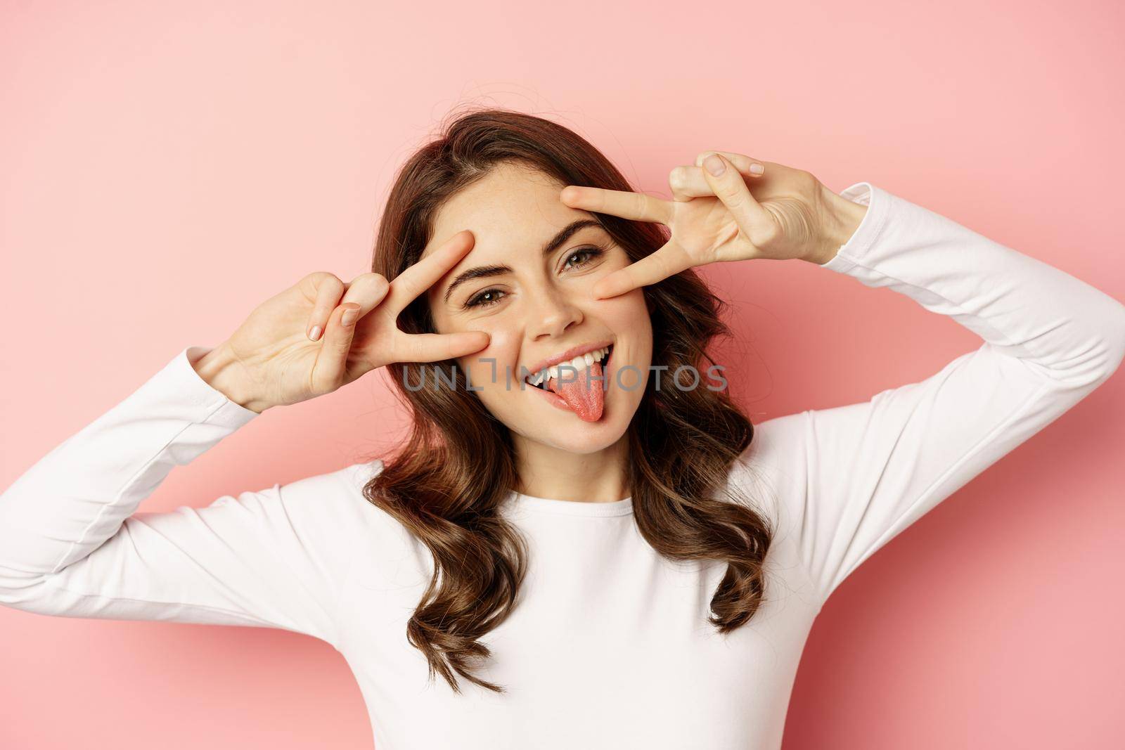 Close up face of young stylish girl, sassy woman showing happy, excited face expression, peace v-sign over eyes, standing against pink background.