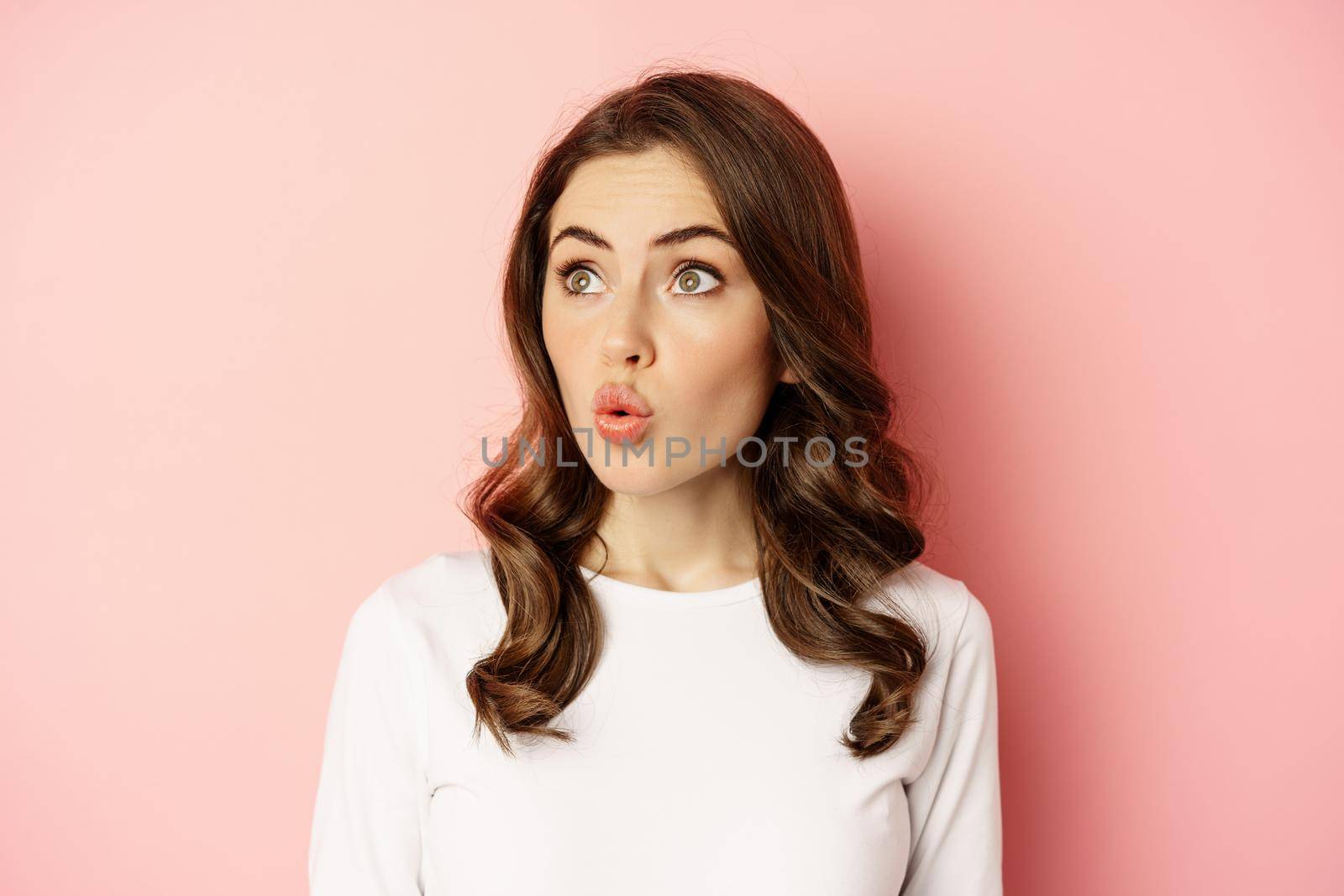 Image of stylish glamour woman looking surprised and amazed, saying wow with impressed reaction, standing over pink background.