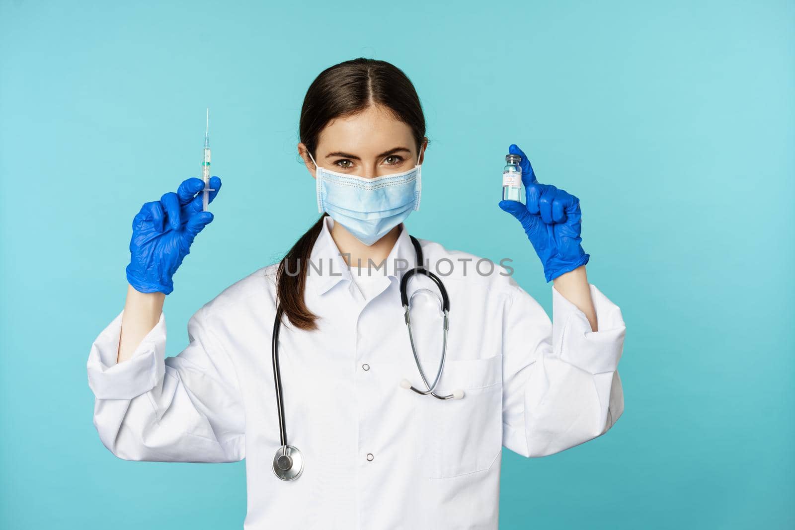 Smiling medical staff, doctor in face mask and rubber gloves, showing syringe and vaccine from covid-19 pandemic, standing over blue background.