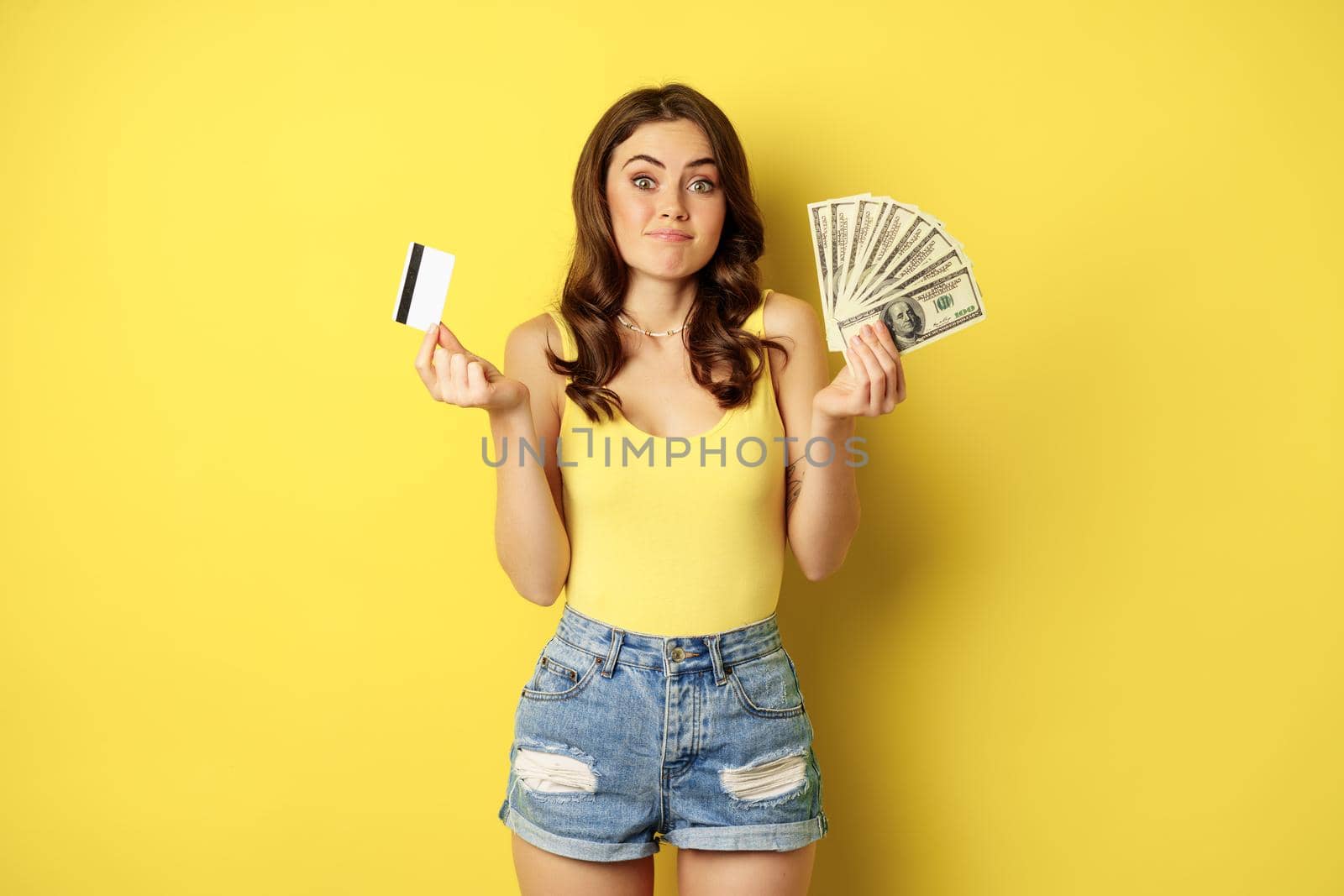 Young pretty woman in summer outfit holding credit card and money, cash in hands, standing against yellow background.