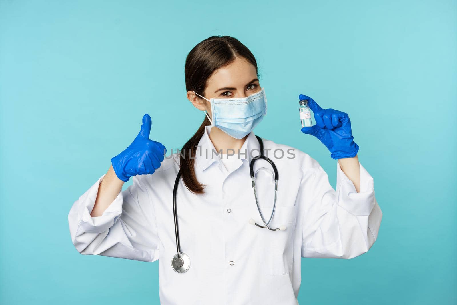 Portrait of smiling doctor, medical personel in face medical mask and rubber gloves, showing thumbs up and coronavirus, omicron vaccine, standing over blue background.