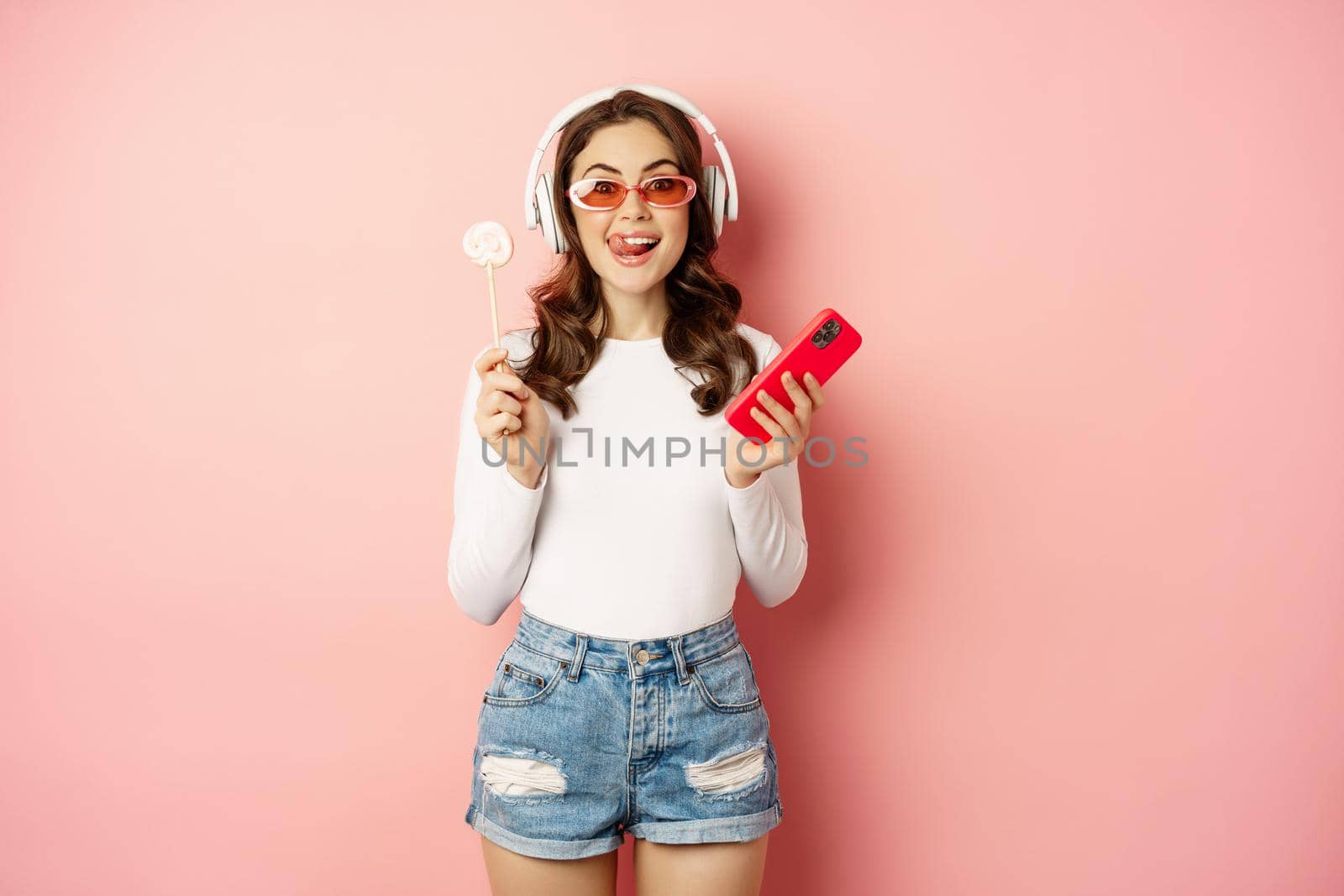 Stylish brunette woman dancing in headphones with mobile phone, listening music in earphones, licking lolipop, standing over pink background by Benzoix