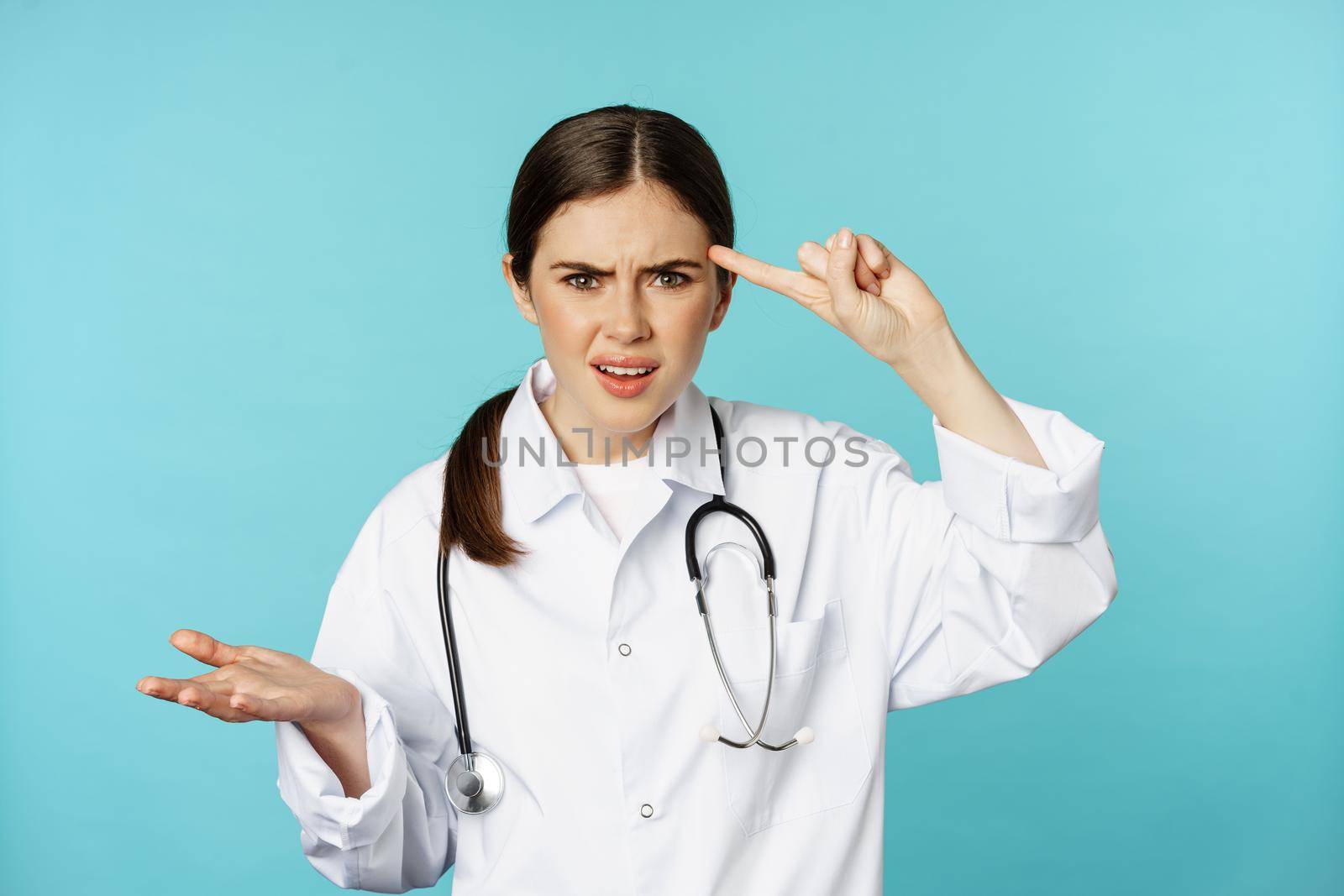 Annoyed woman doctor pointing finger at head, scolding someone stupid, crazy or strange, standing over torquoise background.