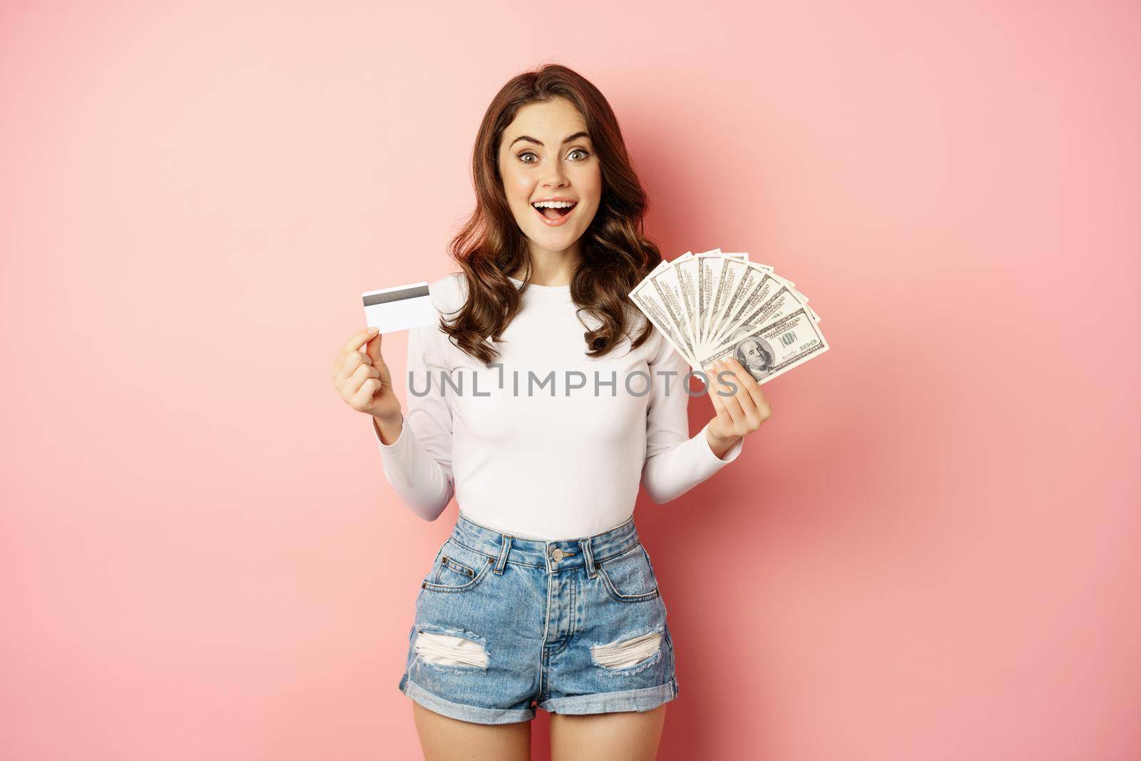 Credit, money and loans concept. Happy beautiful girl holding credit card and cash, looking satisfied, thinking of shopping, standing over pink background by Benzoix
