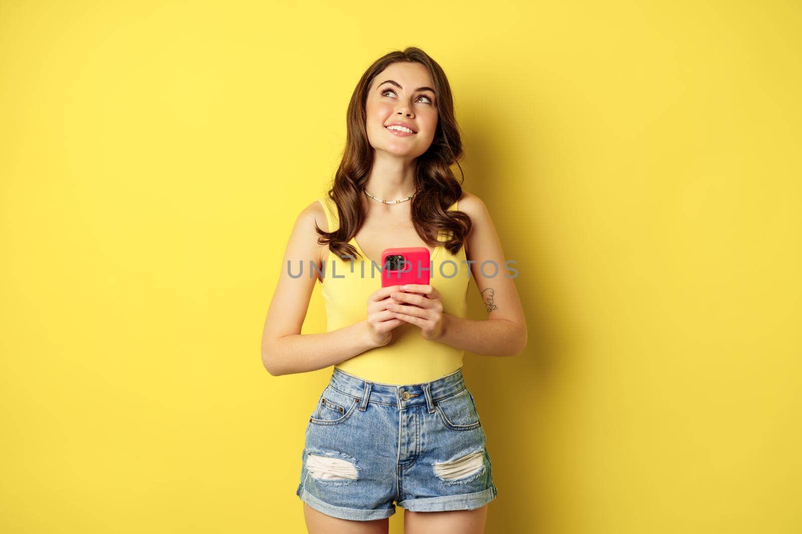 Technology and cellphone. Beautiful stylish girl order in online store, praying in application, using smartphone app and thinking, making decision, standing over yellow background by Benzoix