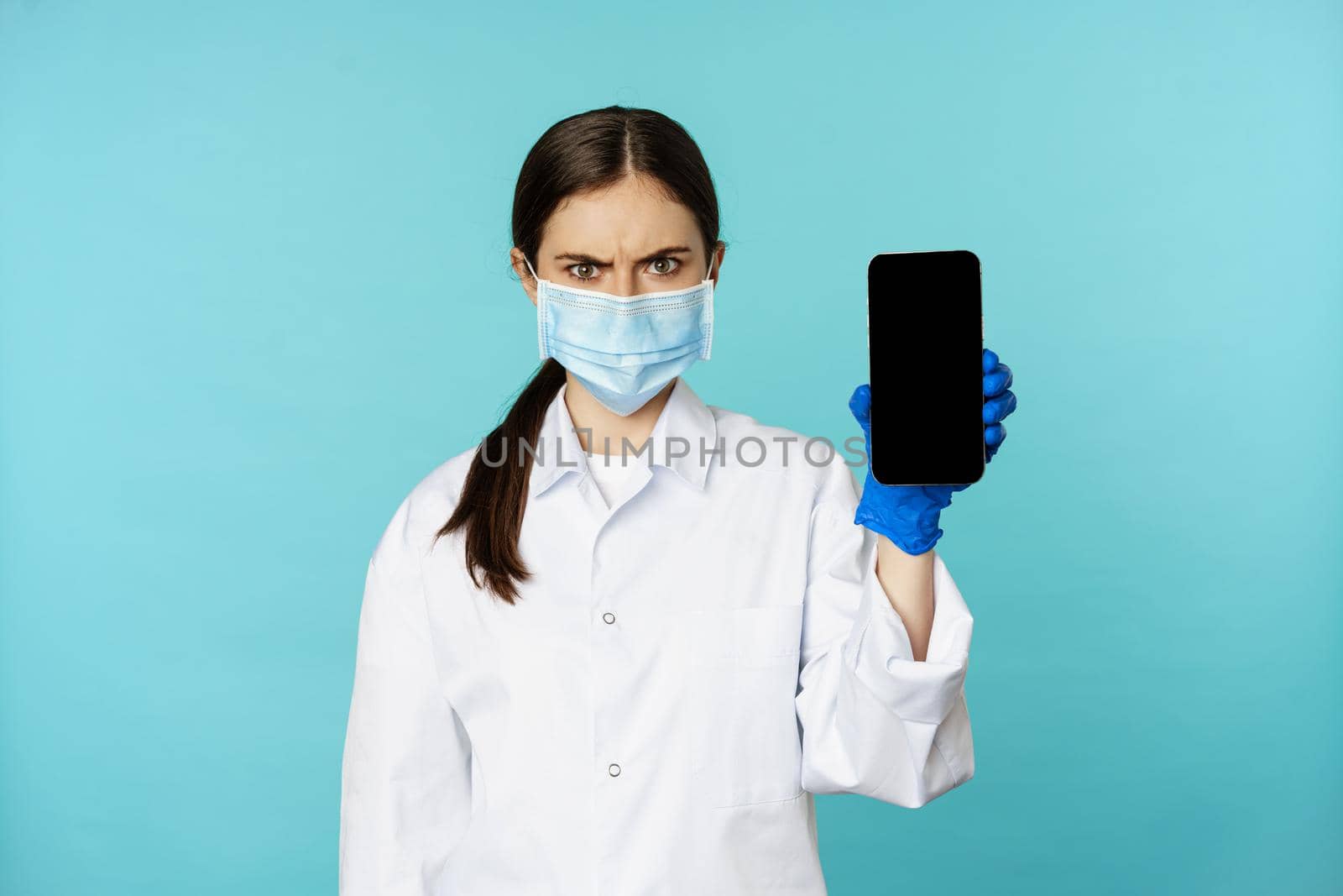 Angry and confused woman doctor in face mask, gloves, showing smartphone app, mobile screen, frowning upset, standing over blue background.