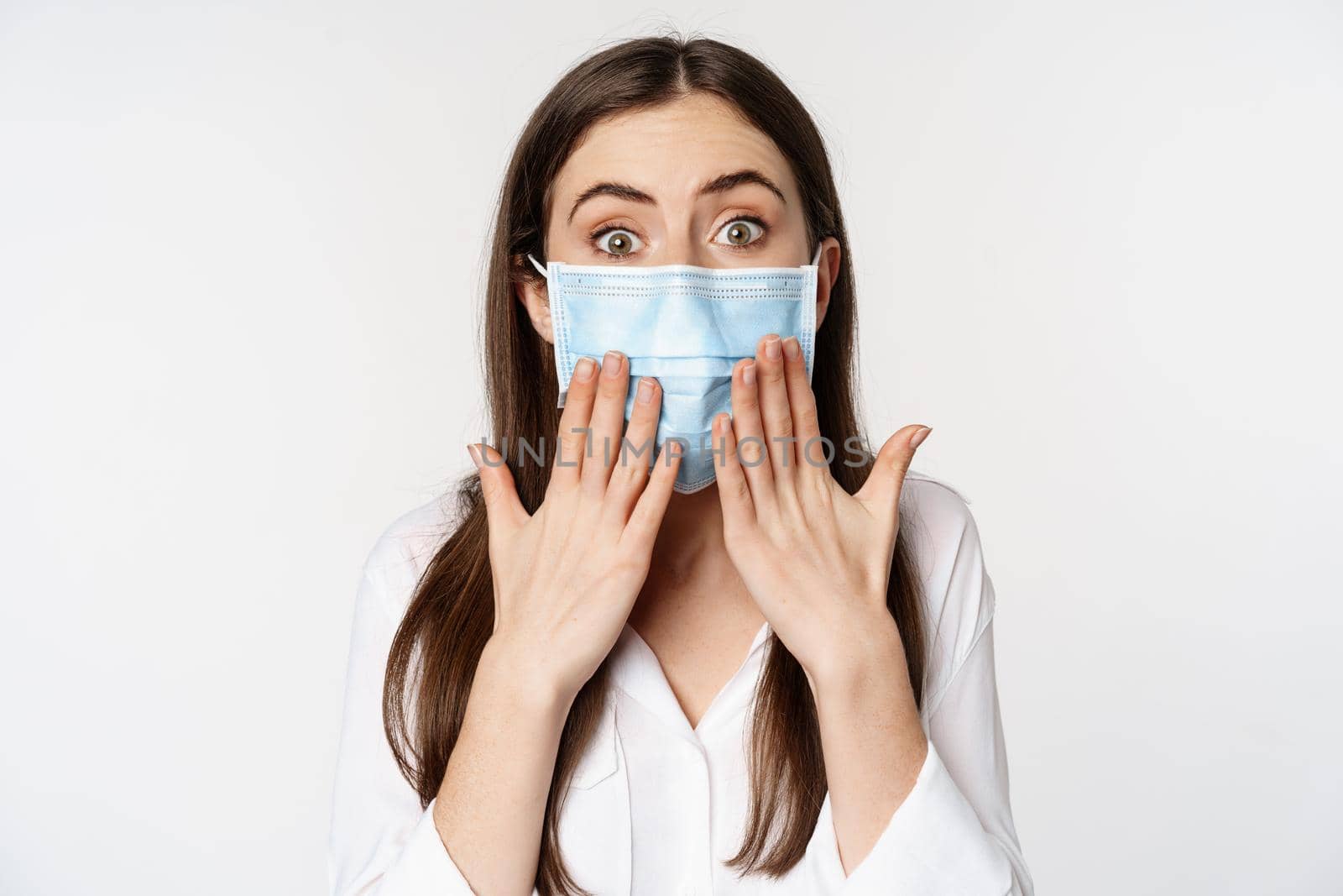 Covid and quarantine gesture. Young woman in face medical mask, looking surprised and shocked at camera, standing amazed against white background.