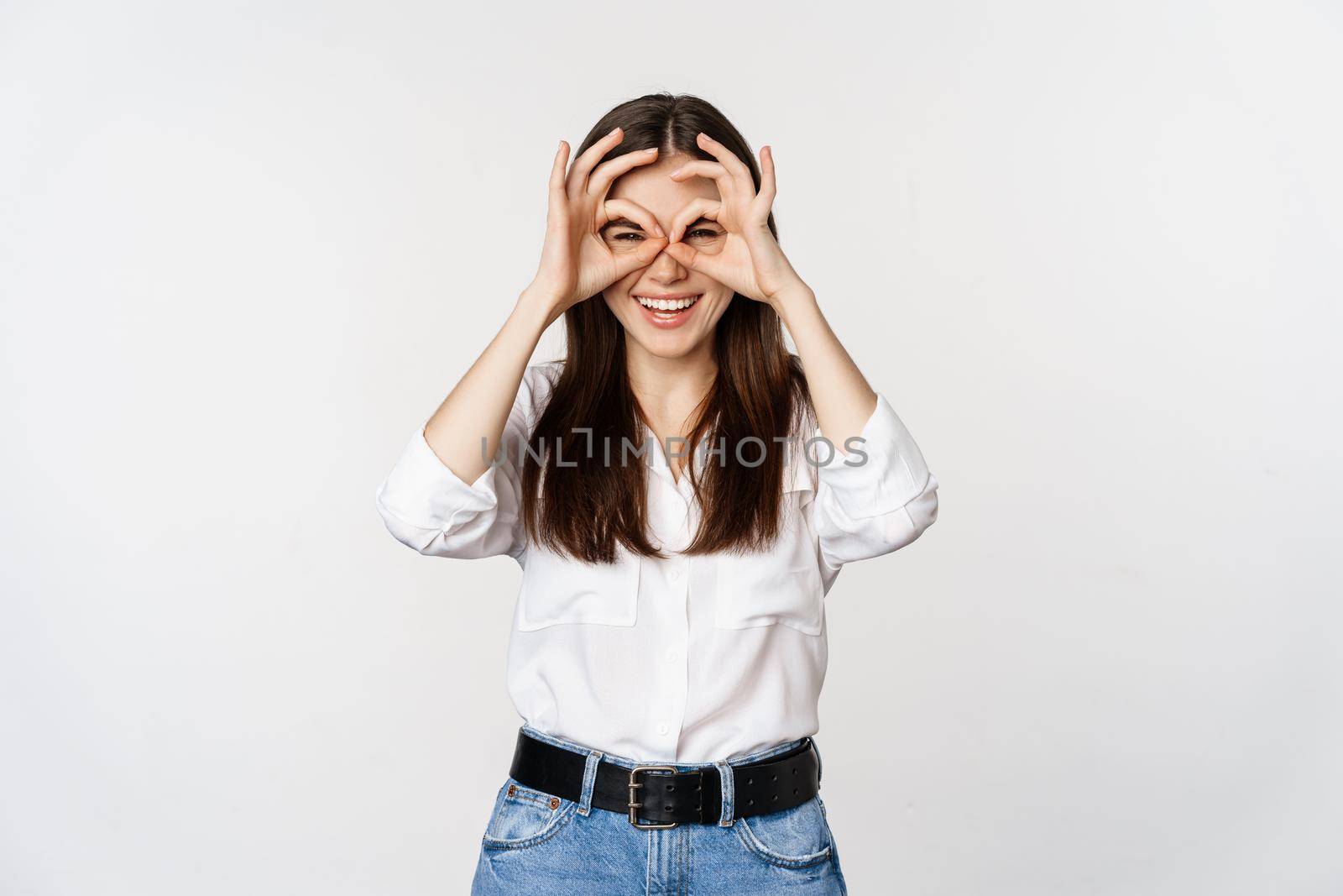 Funny girl looking through hand binoculars, zero gesture, standing over white background and smiling, seeing smth interesting far away, white background.