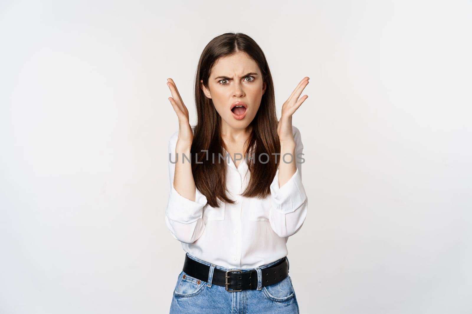 Frustrated and angry young woman gasping, looking shocked and upset at camera, offended by smth, standing bothered against white background.