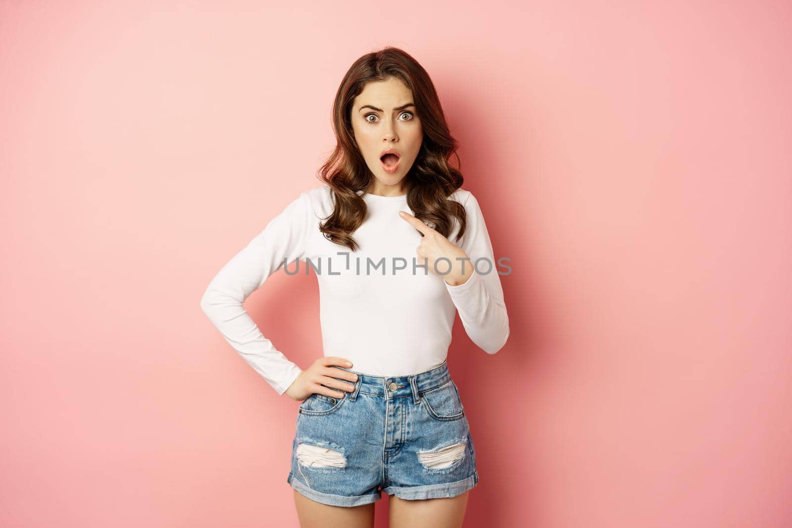 Shocked and offended young beautiful woman, pointing finger at herself, oh my gosh, who me gesture, being insulted, standing in disbelief over pink background.