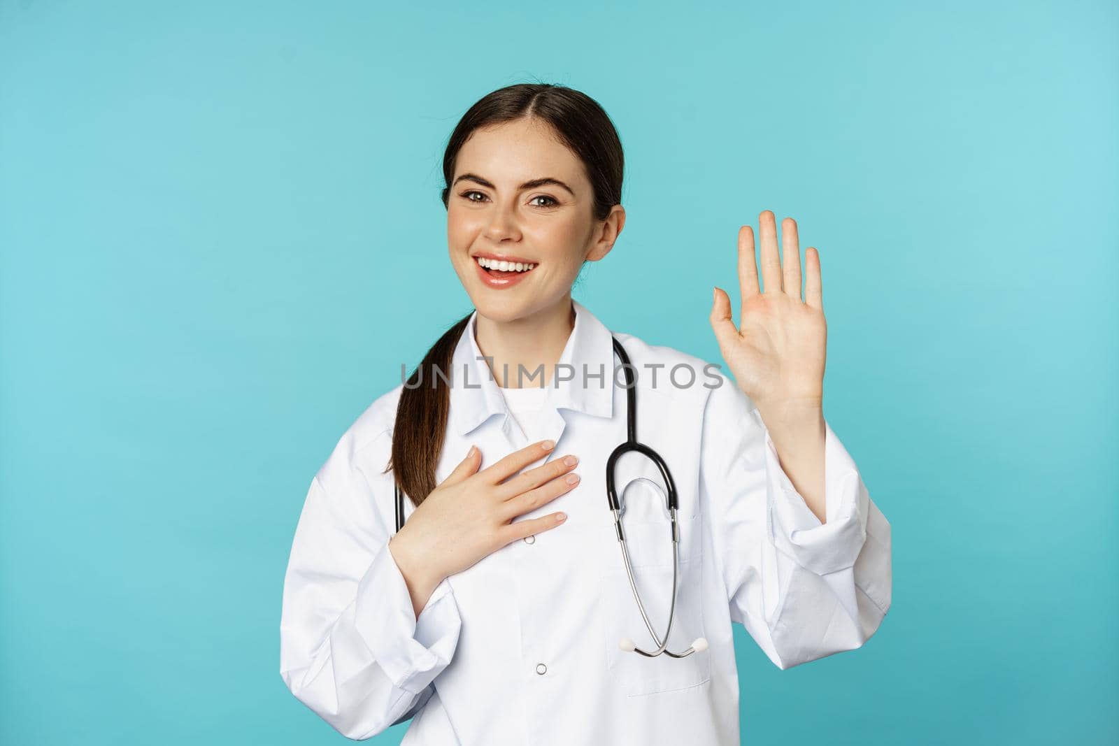 Friendly smiling woman doctor raising hand, name herself, introducing, standing in lab white coat against torquoise background.