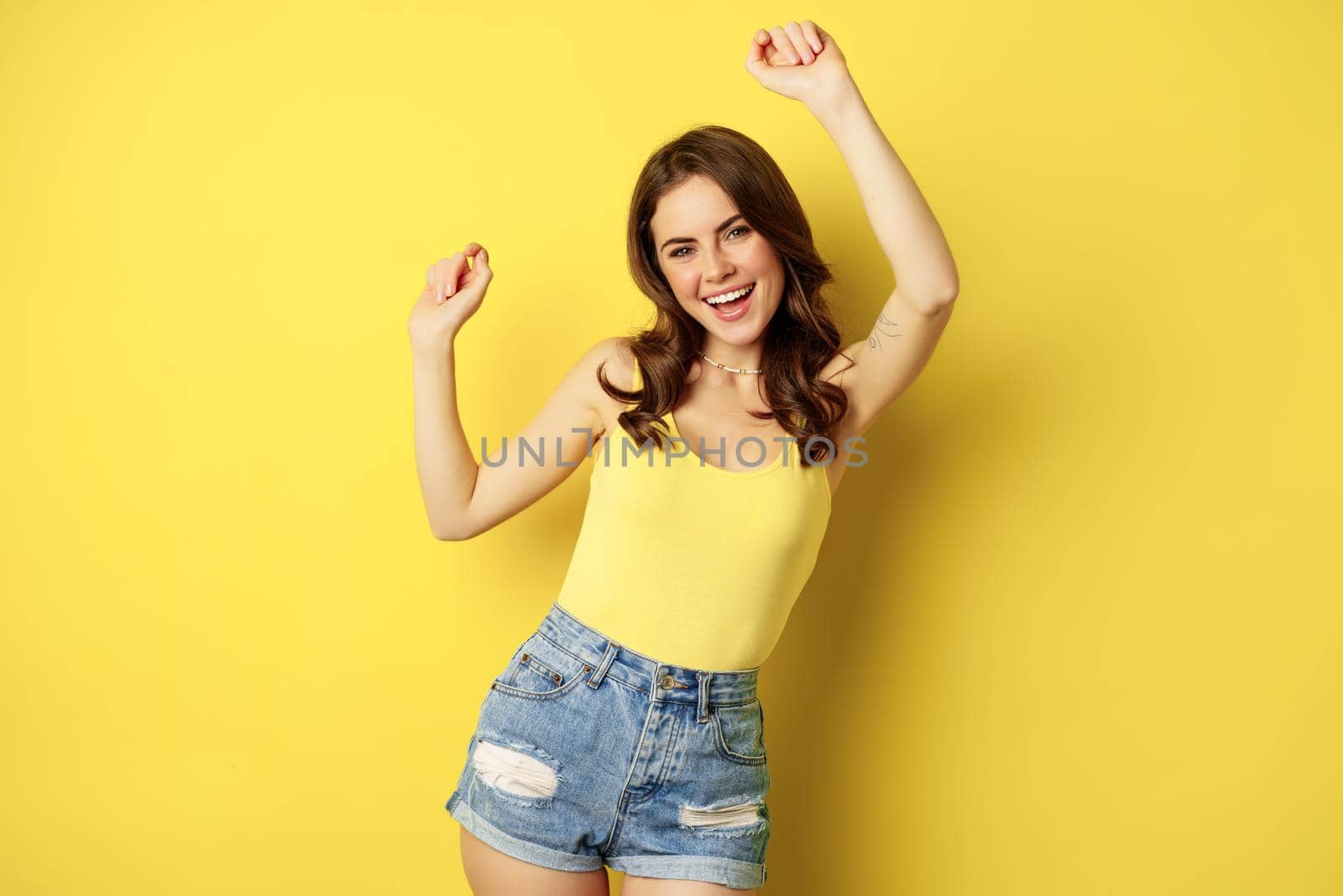 Dancing happy summer girl having fun, raising hands up and rejoicing, standing in tank top and shorts against yellow background by Benzoix