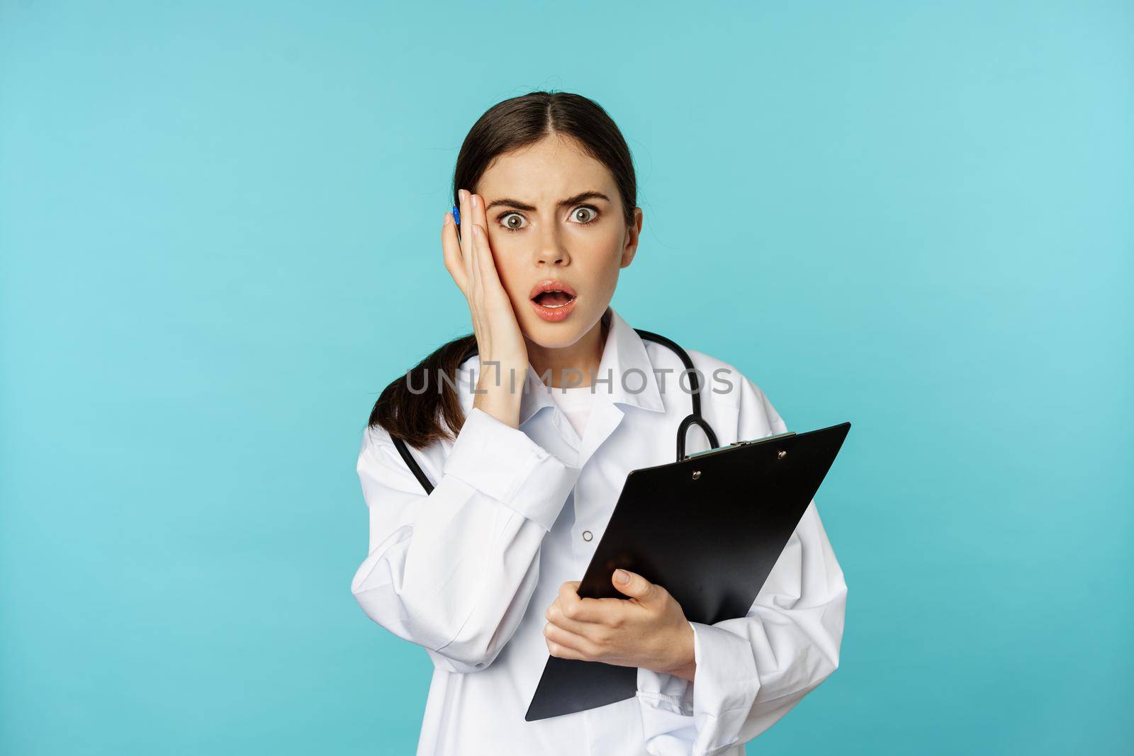 Shocked woman doctor reacting worried and frustrated at camera, hear smth strange, standing in white coat against torquoise background.