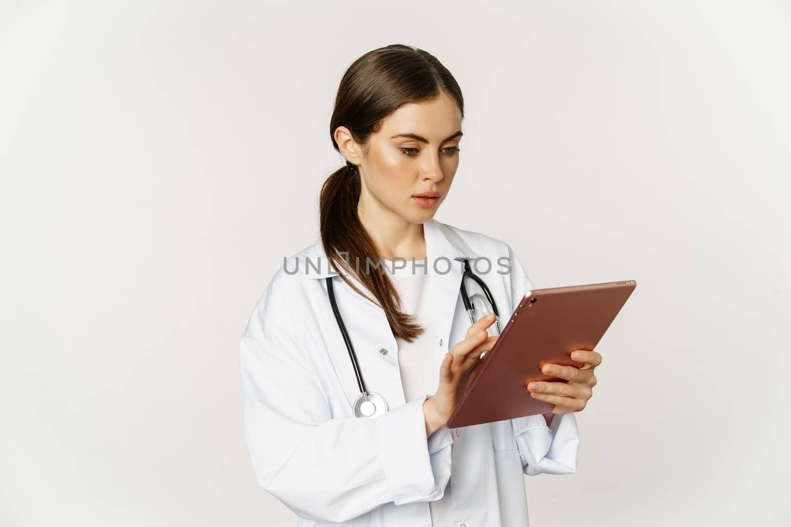 Woman doctor looking concerned at digital tablet, reading with worried face expression, wearing white coat, standing over white background.