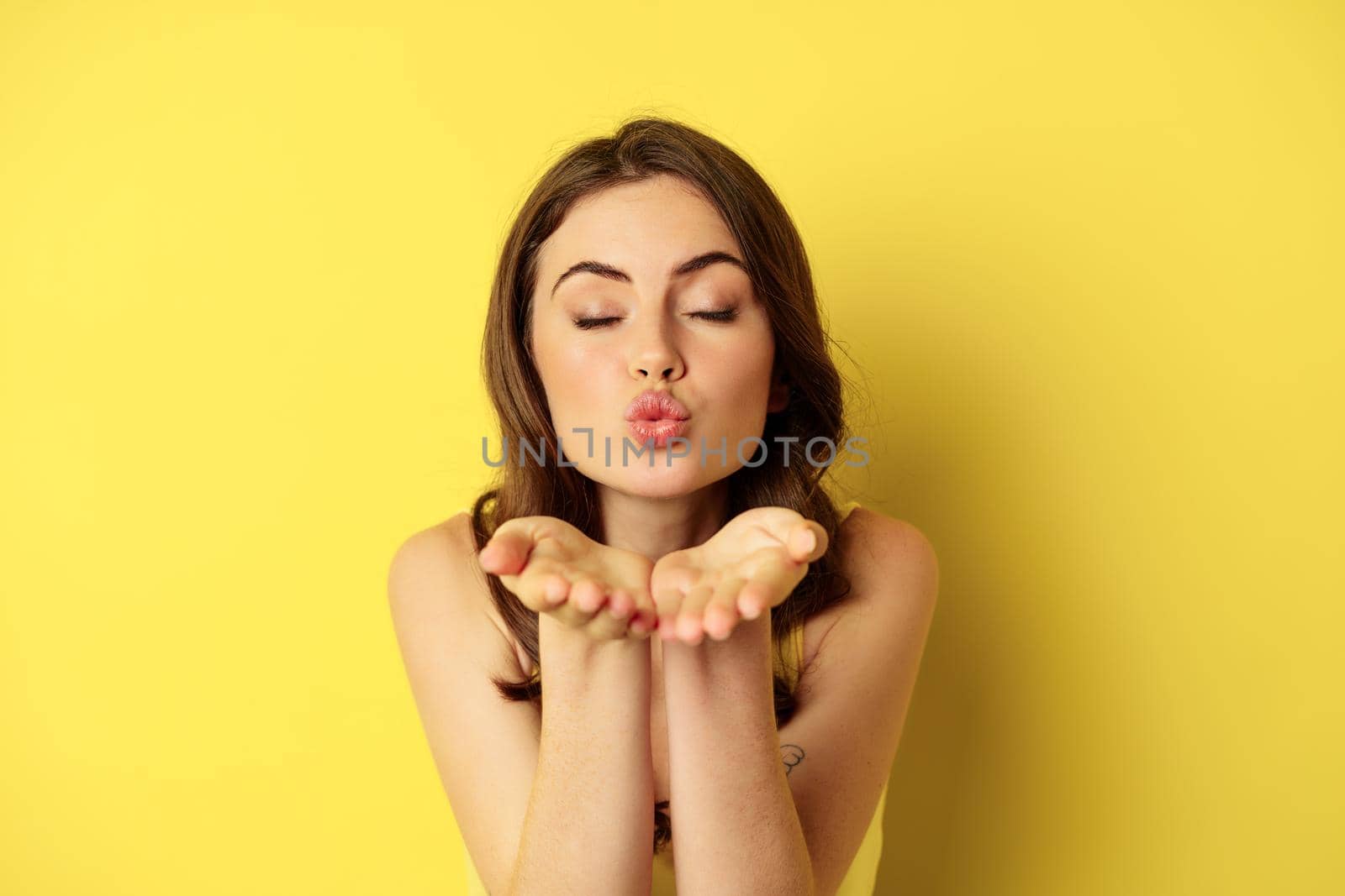 Close up portrait of beautiful, coquettish woman sending air kiss, blowing mwah at camera, standing over yellow background. Copy space