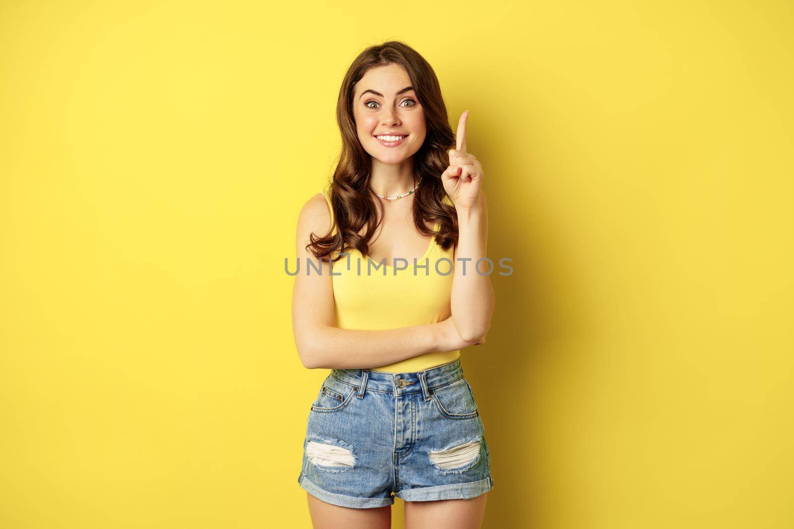 Creative young woman has suggestion, raising finger, eureka sign, has an idea, standing in summer outfit over yellow background. Copy space