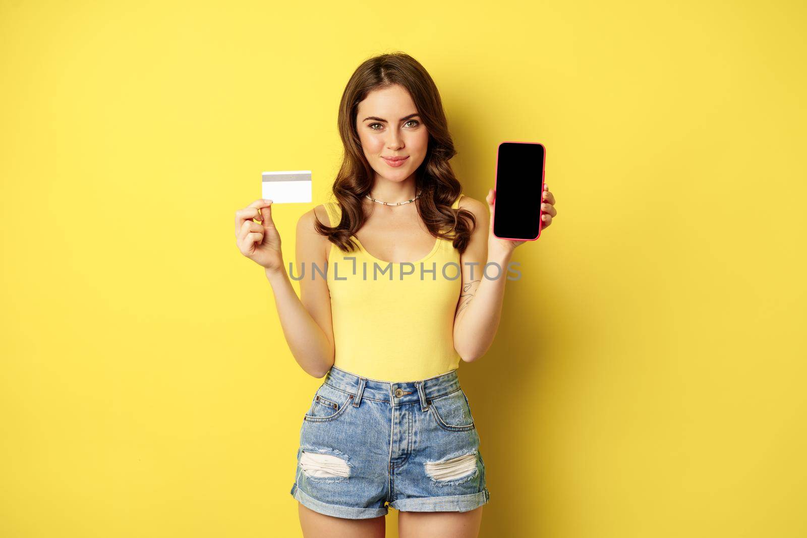 Young beautiful woman model showing smartphone empty phone screen and credit card, ready for summer, wearing tank top and shorts, standing over yellow background.