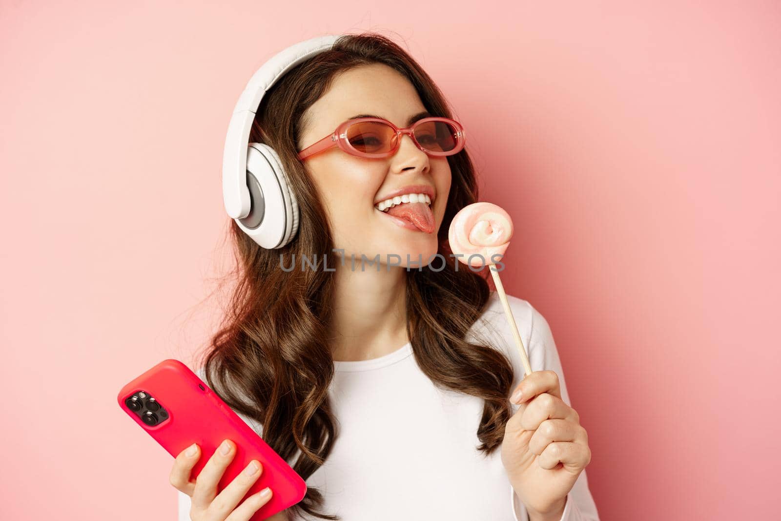 Beautiful female model listening music in headphones, holding lolipop and mobile phone, posing in sunglasses, standing over pink background.