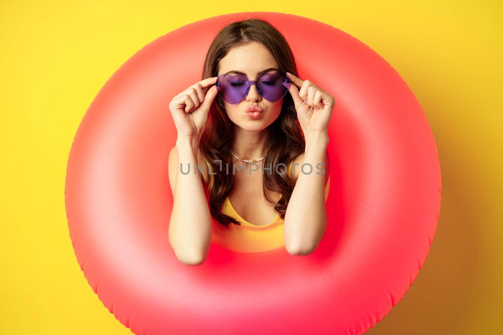 Close up portrait of stylish young woman, girl with swimming ring and summer sunglasses, ready for beach holiday, vacation, standing over yellow background.