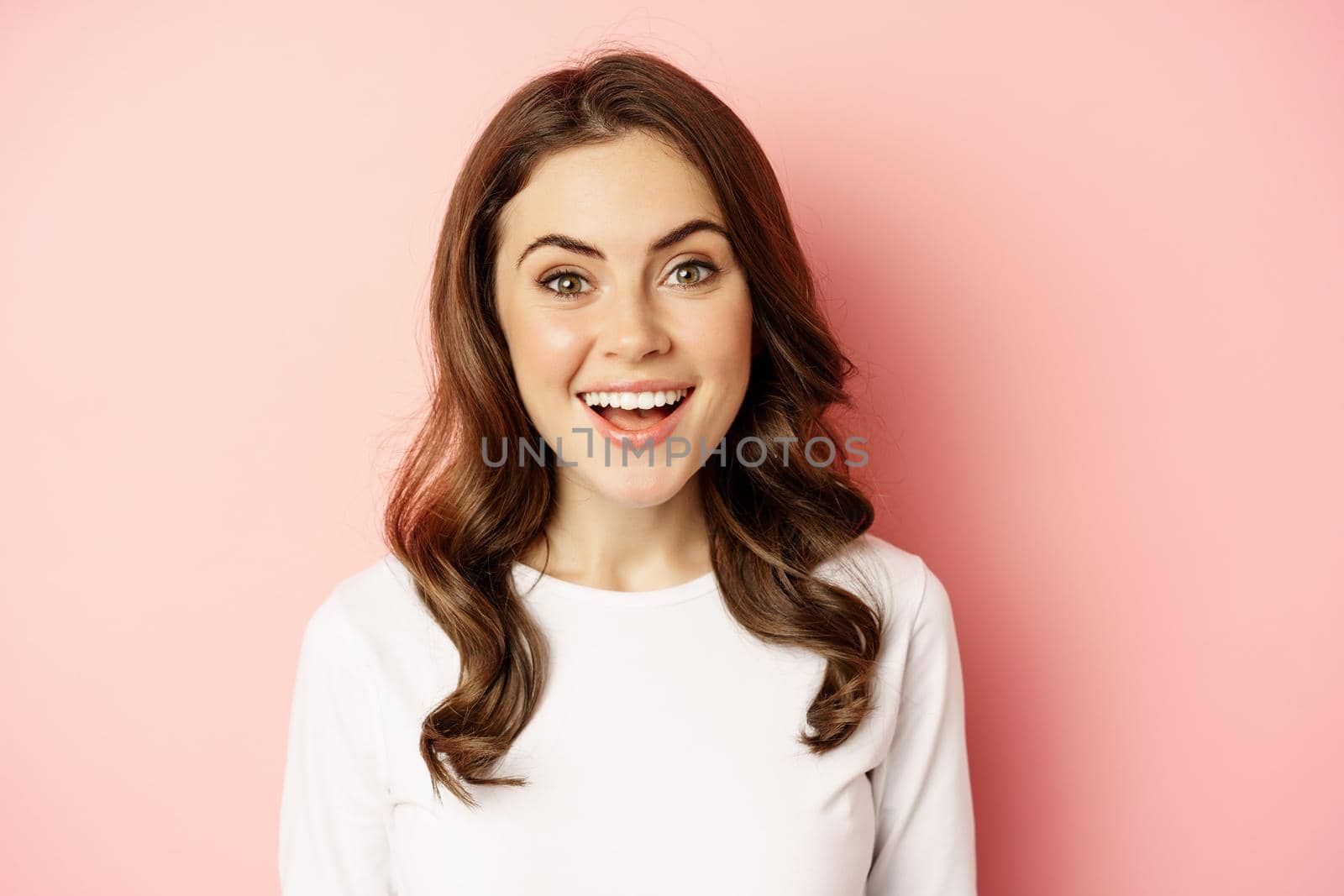 Close up of enthusiastic brunette girl with makeup, smiling and looking happy at camera, posing against pink background by Benzoix