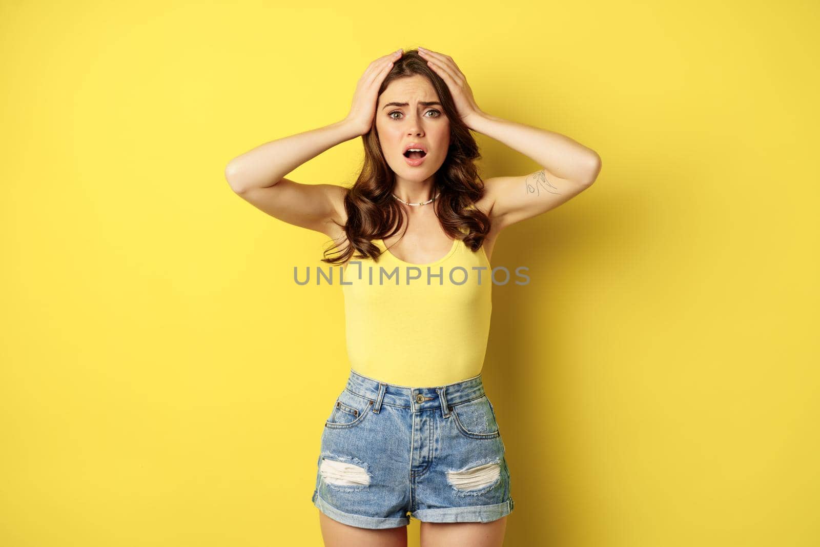 Troubled and shocked woman holding hands on head, panicking, feeling distressed and anxious, having problem, standing over yellow background.