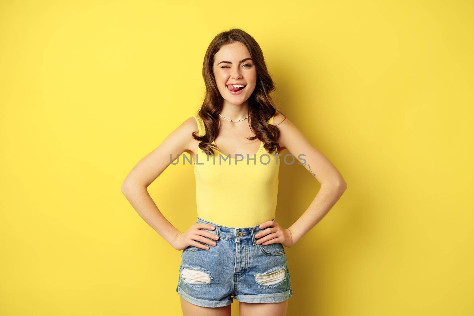 Stylish young brunette woman showing her white healthy teeth, smiling and showing tongue, winking enthusiastic, standing over yellow background.