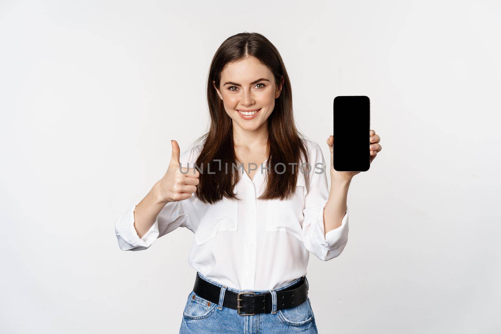 Smiling woman showing thumbs up, recommending app, mobile phone screen, empty interface on smartphone, standing against whte background.