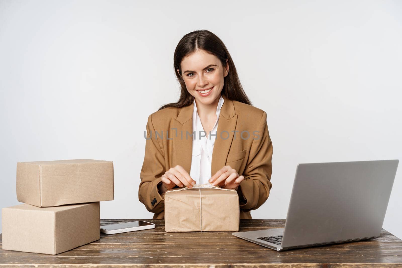 Young businesswoman, self-employed woman wrapping packages, packing clients orders, sitting near laptop and working, white background.