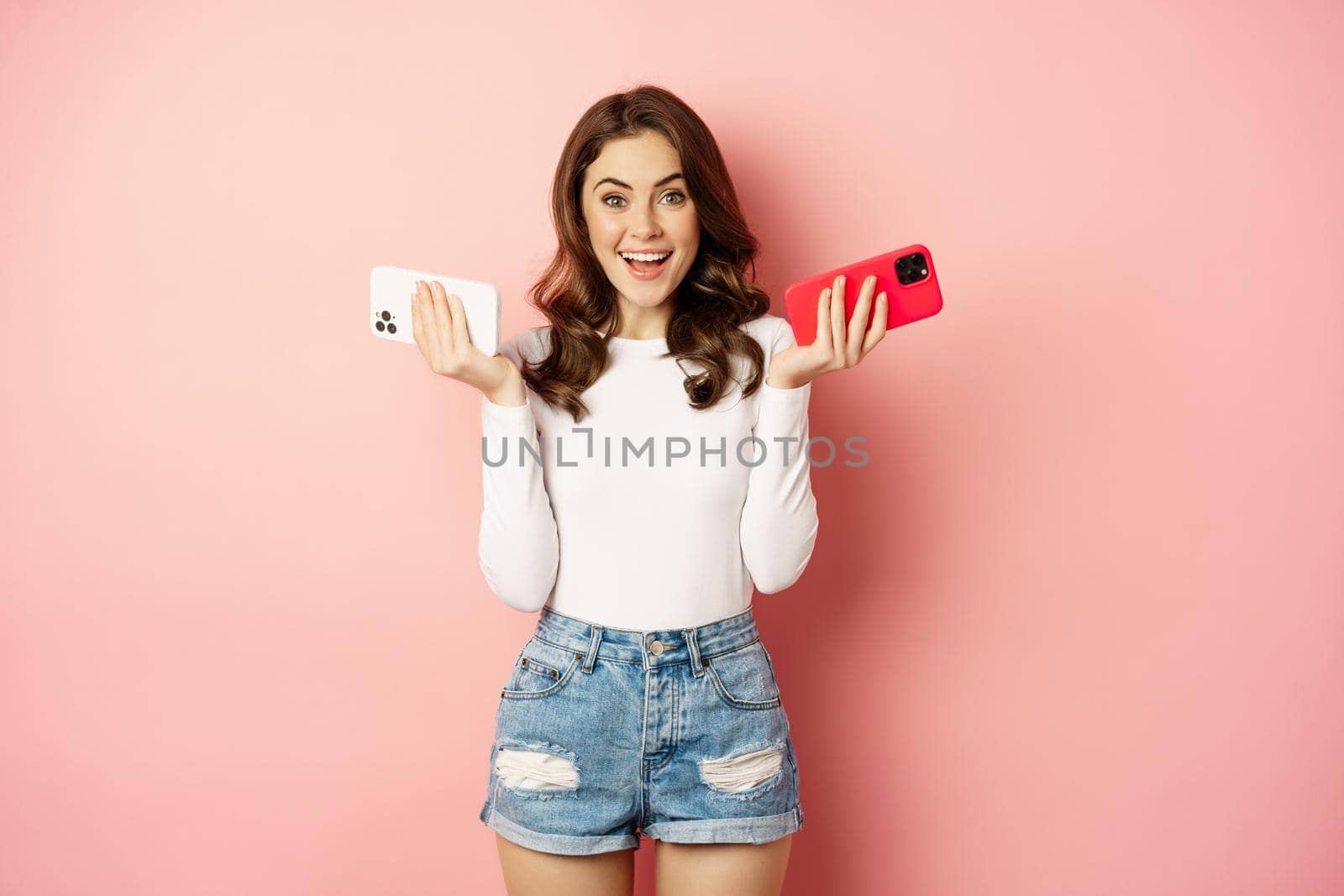 Technology, spring promo concept. Stylish glamour woman holding two smartphones, mobile phones in both hands, smiling pleased, buying new cellphone, pink background by Benzoix
