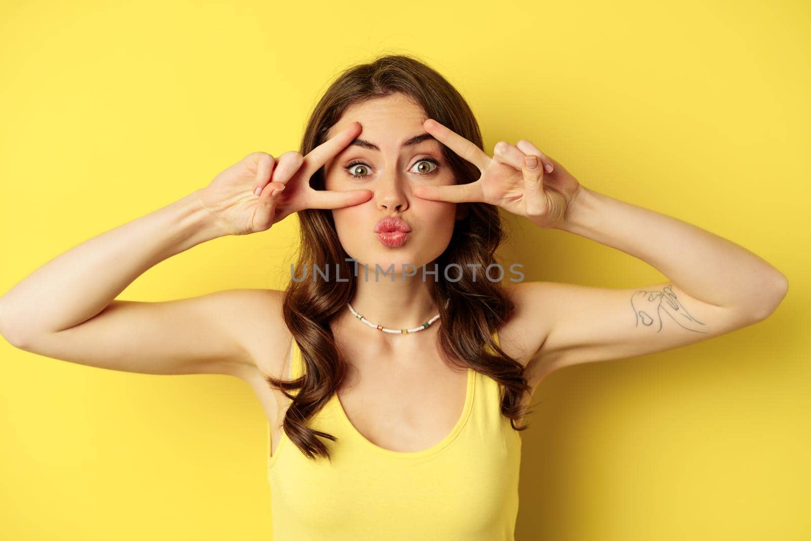 Close up portrait of smiling beautiful girl, showing peace v-sign and looking happy, posing for summer photo, standing against yellow background.