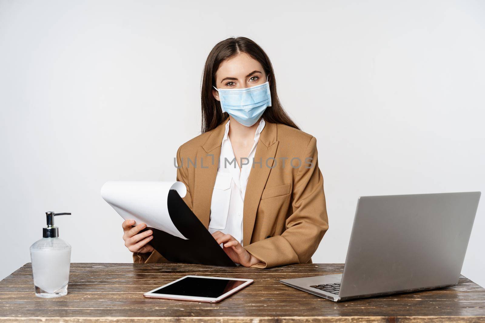 Smiling lady boss, female office worker at workplace, wearing face mask, working with papers and documents at her work desk, white background.