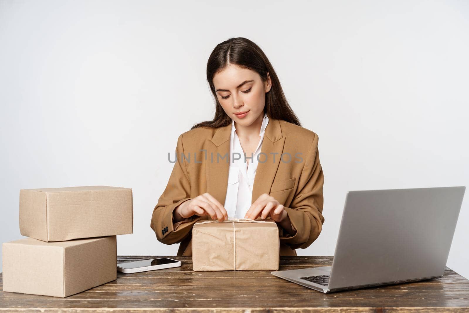 Young businesswoman, self-employed woman sitting in office near laptop, wrapping packages, packing clients orders, working, white background.