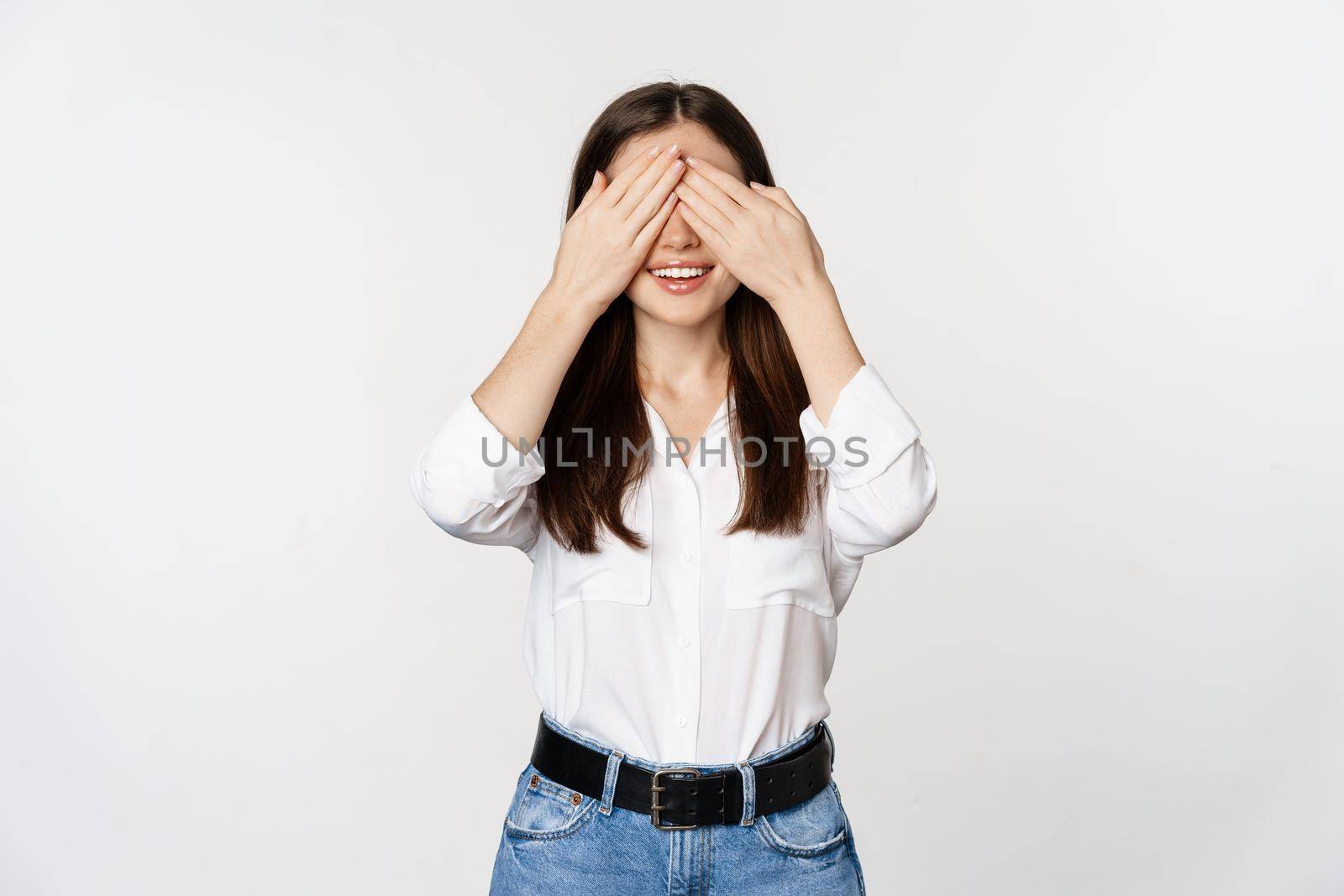 Smiling happy woman waiting for surprise, shut eyes with hands, standing blindsided against white background.