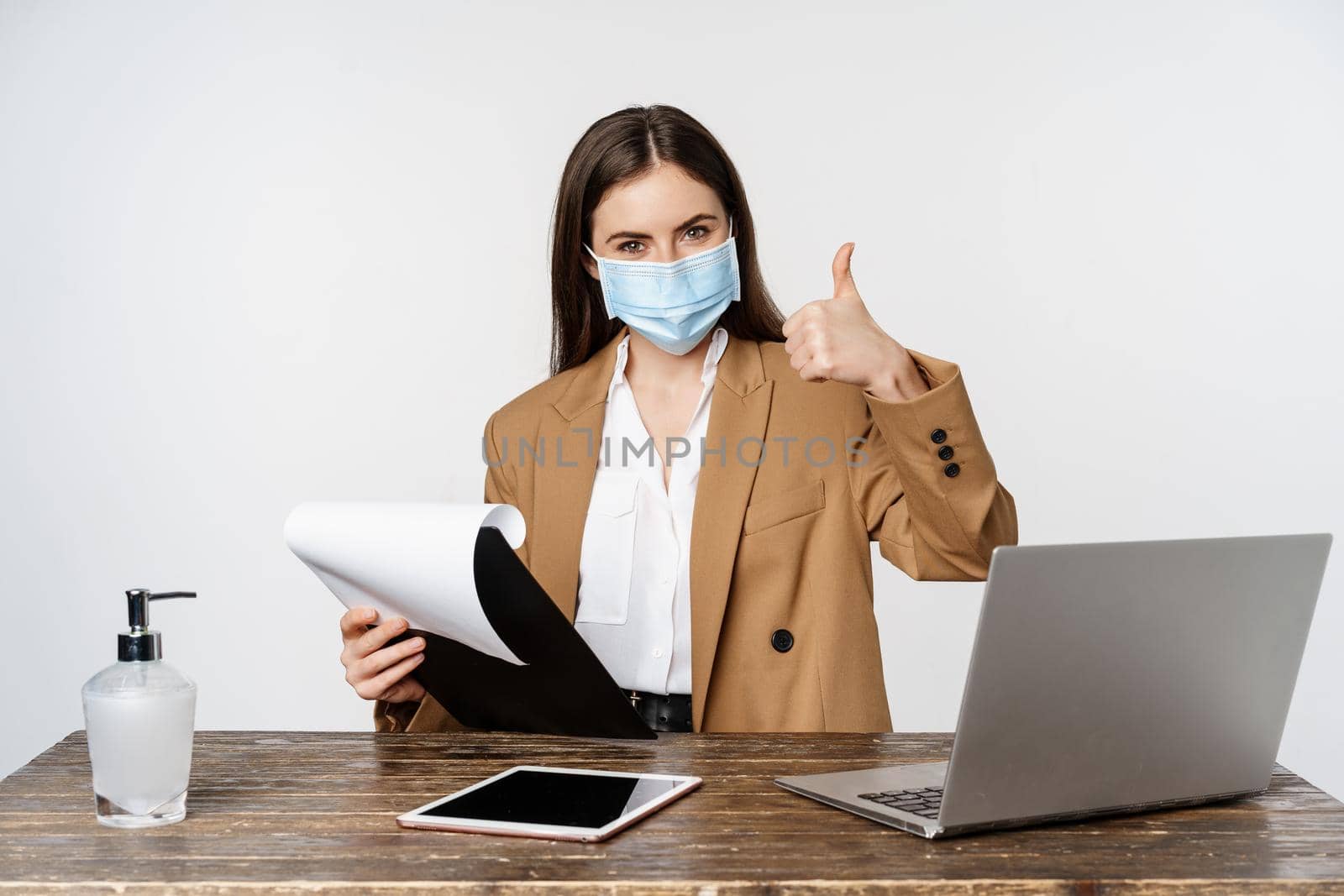 Woman working in office at table, holding clipboard with papers, showing thumbs up, wearing medical face mask, white background.