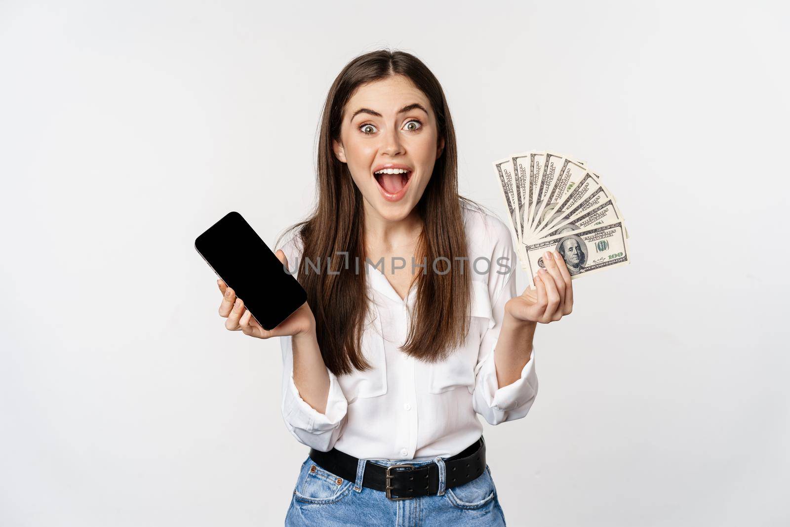 Enthusiastic young woman winning money, showing smartphone app interface and cash, microcredit, prize concept, standing over white background.