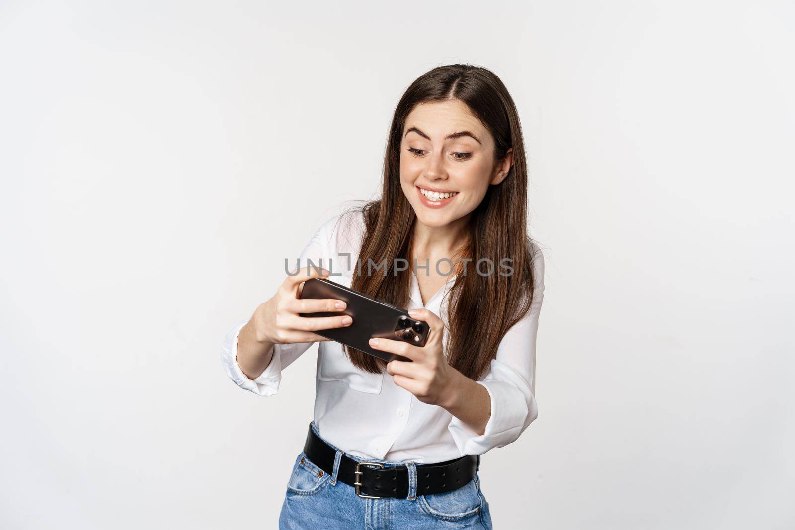 Happy brunette woman playing mobile video game, smiling and looking at screen excited, standing over white background.