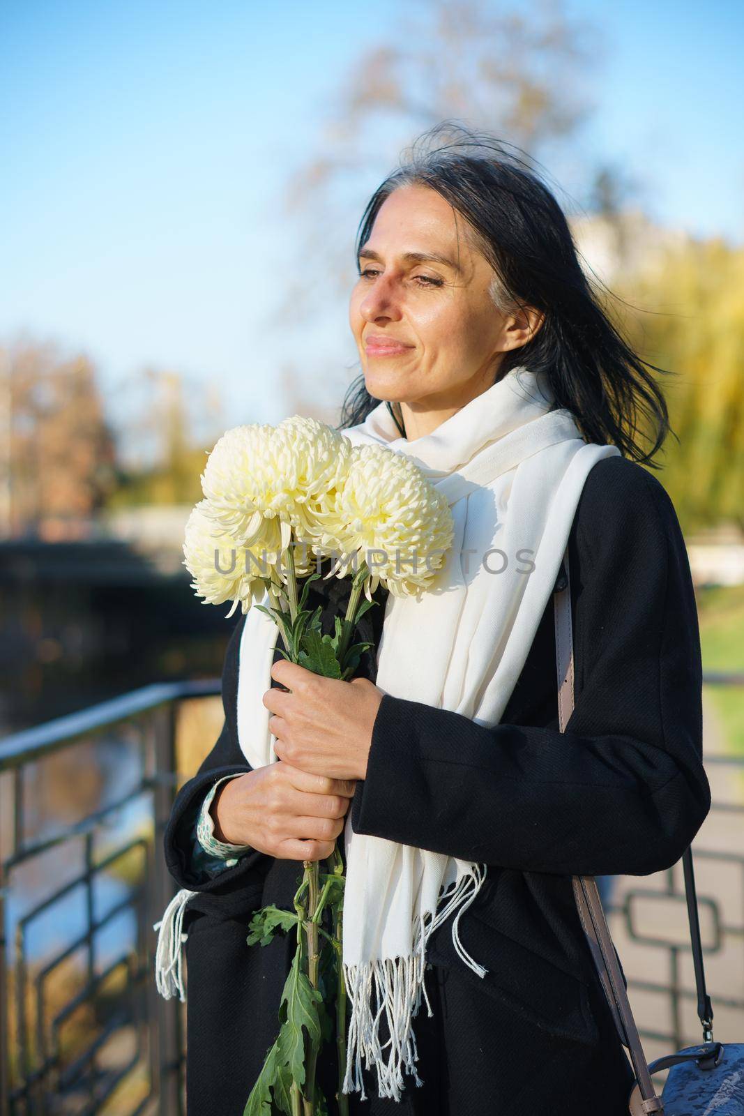A beautiful middle-aged woman getting grey-haired in a dark coat in a spring town with a bouquet of flowers. by balinska_lv
